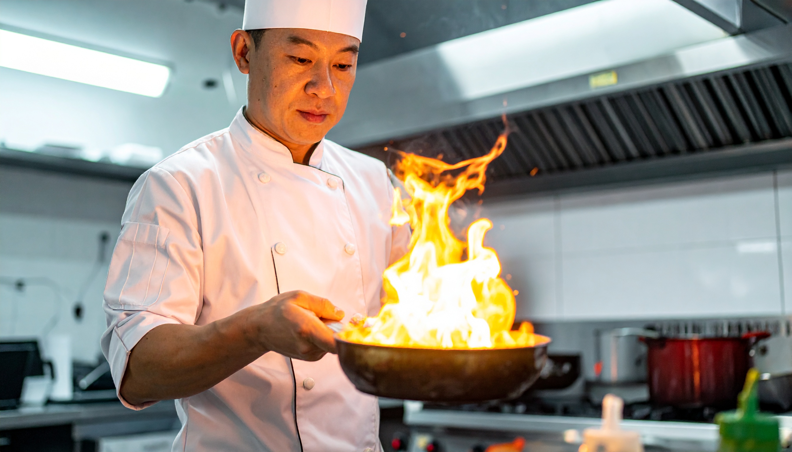 Chef de cozinha em uniforme branco prepara um prato flamejante em uma frigideira, destacando-se contra o fundo profissional da cozinha. A iluminação suave realça a intensidade das chamas, criando um contraste dramático e focando na expressão concentrada do chef. O ambiente é moderno e bem equipado, sugerindo um espaço culinário de alta qualidade.